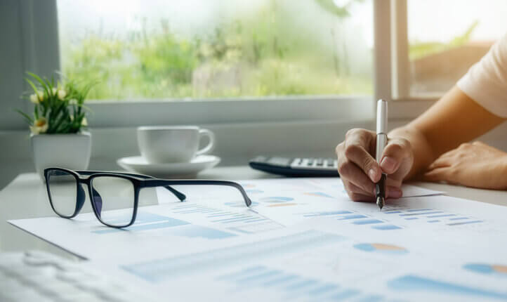 Someone writing notes at a desk using a pen, with glasses, coffee and calculator in view