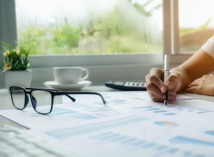 Someone writing notes at a desk using a pen, with glasses, coffee and calculator in view
