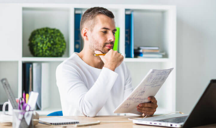 young man looking at paperwork at his desk, in front of his laptop