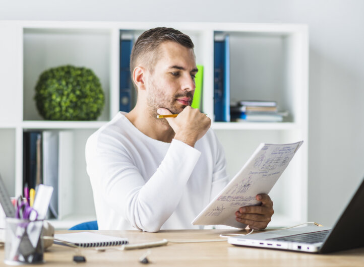 young man looking at paperwork at his desk, in front of his laptop